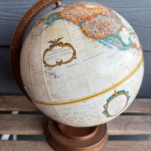 Może przedstawiać: Zabytkowy globus Replogle World Classic Series o średnicy 9 cali. Globus ma kremową powierzchnię ze szczegółową mapą świata, zaakcentowaną brązowymi i złotymi tonami. Jest zamontowany na podstawie w kolorze brązu i ma brązowy pierścień południka.