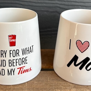 May include: Two white ceramic mugs with black and red text. One mug says "Sorry for what I said before I had my Tims" and the other says "I ❤️ You Mom". Both mugs have a white handle.