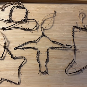 May include: Five rustic metal ornaments shaped like a cowboy hat, star, coat hanger, and cowboy boot. Each ornament is made from dark brown barbed wire and has a twine loop for hanging. The ornaments are displayed on a light brown wooden surface.