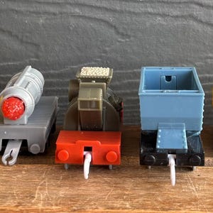 Könnte beinhalten: Eine Sammlung bunter Spielzeug-Zugwaggons, darunter rot, grün, gelb, grau und blau. Die Waggons sind in einer Reihe auf einer Holzoberfläche angeordnet, jedes mit einem einzigartigen Design. Die Spielzeuge sind aus Kunststoff.