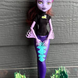 May include: A Monster High doll with a purple mermaid tail and green fins. The doll has purple skin, yellow eyes, and dark red hair with teal and purple highlights. She wears a black top with a small graphic.