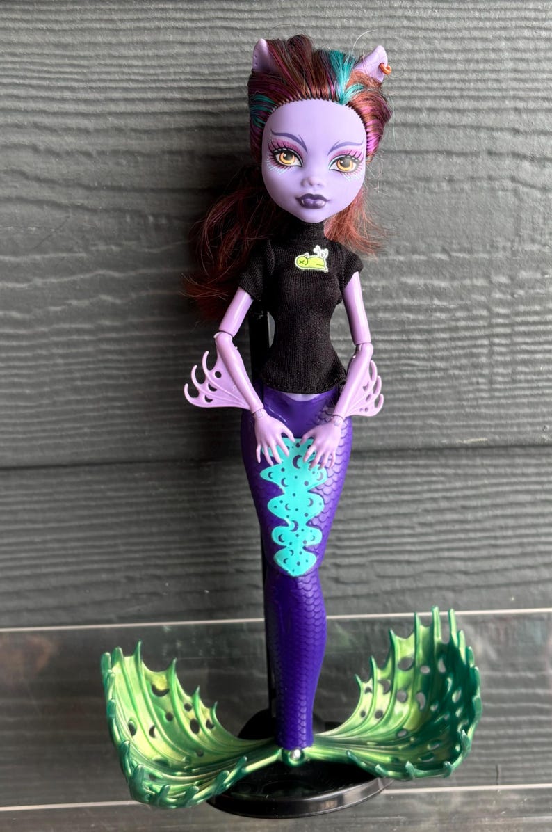 May include: A Monster High doll with a purple mermaid tail and green fins. The doll has purple skin, yellow eyes, and dark red hair with teal and purple highlights. She wears a black top with a small graphic.