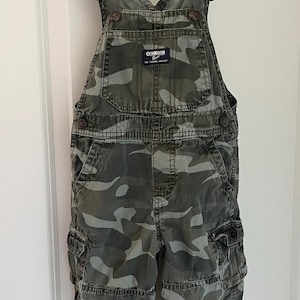 Puede incluir: Pantal&oacute;n de peto de camuflaje para un ni&ntilde;o. El peto es verde y gris con una correa ajustable marr&oacute;n. El peto tiene un parche en la parte delantera que dice "OSHKOSH B'Gosh" y "The Genuine Article".