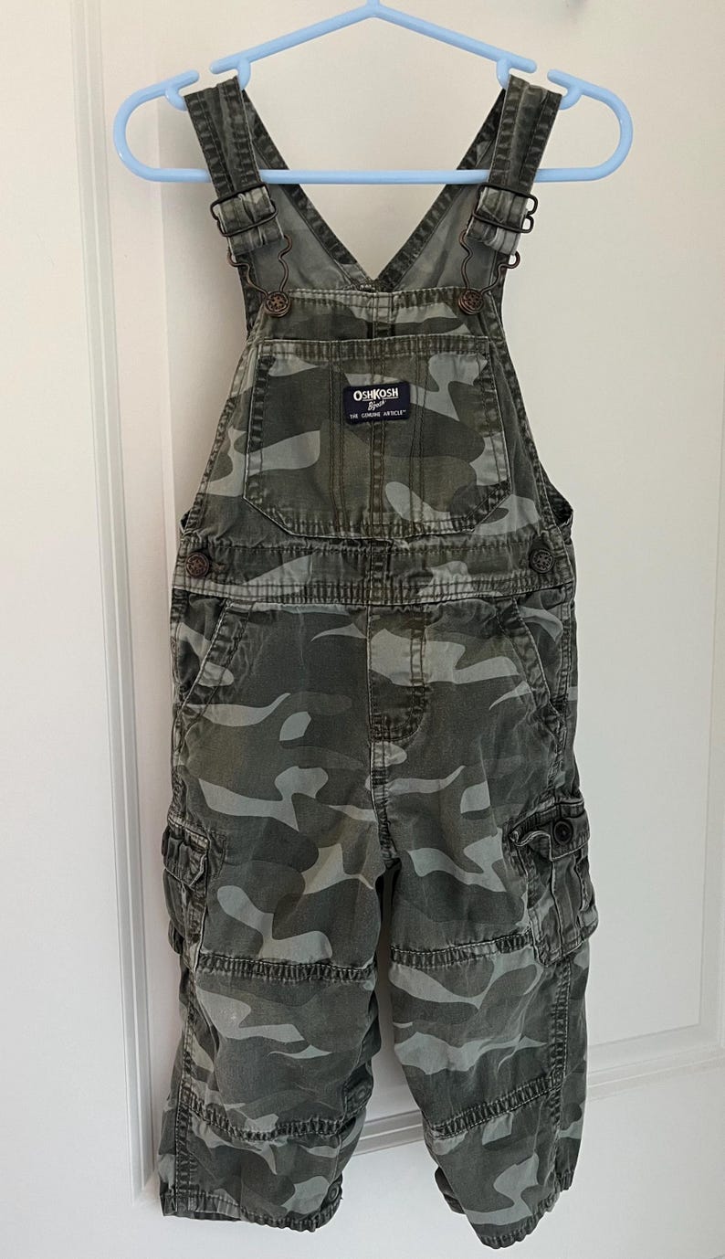 Puede incluir: Pantal&oacute;n de peto de camuflaje para un ni&ntilde;o. El peto es verde y gris con una correa ajustable marr&oacute;n. El peto tiene un parche en la parte delantera que dice "OSHKOSH B'Gosh" y "The Genuine Article".