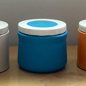 May include: Three round plastic containers with lids. The container on the left is white with a red lid. The container in the middle is blue with a white lid. The container on the right is orange with a white lid.