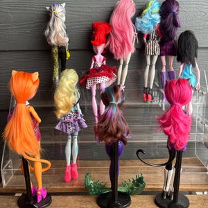 May include: A selection of ten fashion dolls with diverse hair colours and styles, including pink, blue, and purple. The dolls are displayed on a clear acrylic shelf, each with unique outfits and accessories. The dolls are standing on black stands.