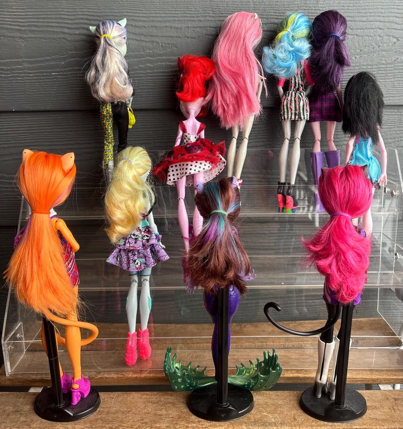 May include: A selection of ten fashion dolls with diverse hair colours and styles, including pink, blue, and purple. The dolls are displayed on a clear acrylic shelf, each with unique outfits and accessories. The dolls are standing on black stands.