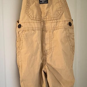 Puede incluir: Pantal&oacute;n vaquero de color beige con forro de cuadros azules y blancos en las tirantes y los pu&ntilde;os. El pantal&oacute;n vaquero tiene un parche en la parte delantera que dice "OshKosh B'Gosh The Genuine Article".