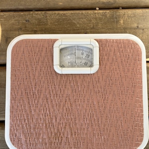 May include: A white bathroom scale with a brown woven platform and a round dial that reads "pounds" and "kilos". The dial has black markings and a white needle. The scale reads "Not Legal for Trade".