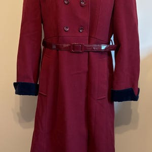 Peut inclure: Un manteau bordeaux à double boutonnage avec une ceinture assortie. Le manteau a un col et des poignets en velours bleu marine. Le manteau est présenté sur un mannequin.