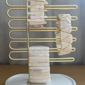 May include: A gold metal stand with a white ceramic base holds two white shell bracelets. The bracelets are made of flat, rectangular pieces of shell.