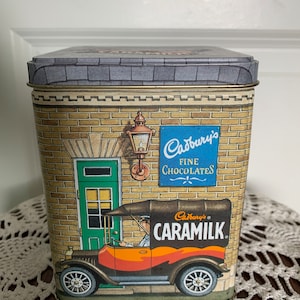 Może przedstawiać: Metalowe pudełko w stylu vintage z ceglanym budynkiem i ciężarówką dostawczą z przodu. Ciężarówka jest pomarańczowa i brązowa z napisem "Cadbury's Caramilk" z boku. Budynek ma znak z napisem "Cadbury's Fine Chocolates".