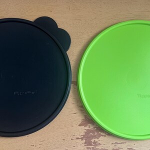 May include: Two round plastic lids, one black and one green. Both lids have a raised lip around the edge and a small knob on the top. The black lid has the word "Tupperware" printed on it, and the green lid has the word "Tupperware" printed on it.