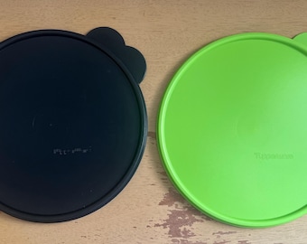 May include: Two round plastic lids, one black and one green. Both lids have a raised lip around the edge and a small knob on the top. The black lid has the word "Tupperware" printed on it, and the green lid has the word "Tupperware" printed on it.
