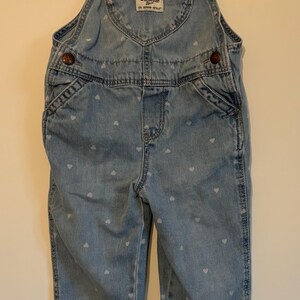 K&ouml;nnte beinhalten: Hellblaue Jeans-Latzhose mit wei&szlig;em Herzmuster. Mit herzf&ouml;rmiger Tasche und dem Logo "OSHKOSH B'GOSH". Verstellbare Tr&auml;ger und Metallbeschl&auml;ge. Ein klassisches Kinderkleidungsst&uuml;ck.