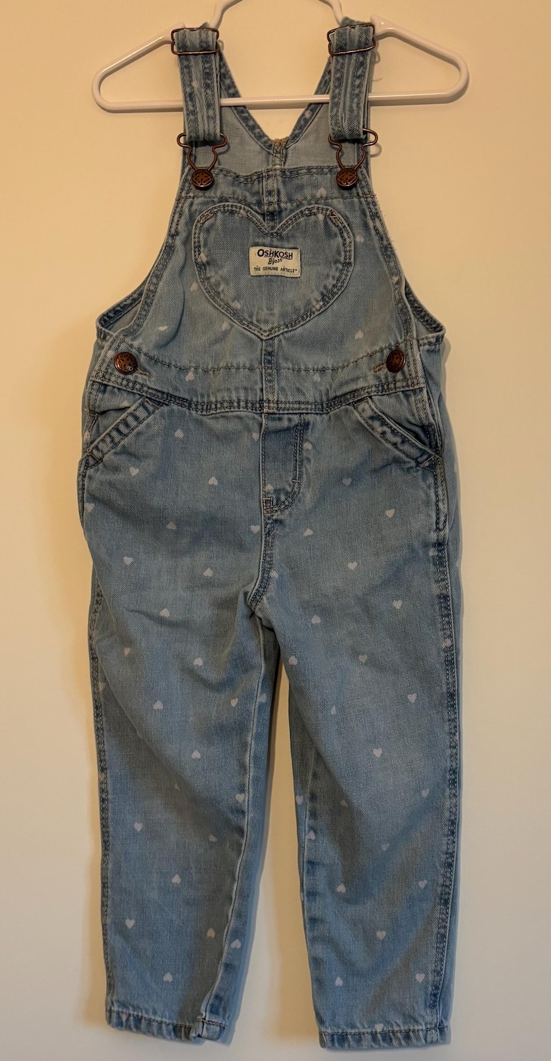 K&ouml;nnte beinhalten: Hellblaue Jeans-Latzhose mit wei&szlig;em Herzmuster. Mit herzf&ouml;rmiger Tasche und dem Logo "OSHKOSH B'GOSH". Verstellbare Tr&auml;ger und Metallbeschl&auml;ge. Ein klassisches Kinderkleidungsst&uuml;ck.