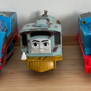 Può includere: Tre locomotive giocattolo in fila. I treni sono blu, rossi e verde acqua con facce sorridenti. Il treno a sinistra è blu con un frontale nero. Il treno centrale è verde acqua con un frontale dorato. Il treno a destra è blu con una base rossa.