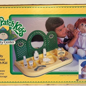 Puede incluir: Caja del set de juego Cabbage Patch Kids Vanity Center. La caja es amarilla con un borde verde y presenta a un niño jugando con una muñeca Cabbage Patch Kid. El set incluye un tocador con espejo y piezas de juego.