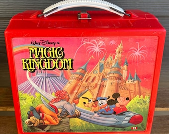 May include: Red plastic lunchbox featuring a vintage illustration of Walt Disney's Magic Kingdom. The image includes Cinderella's Castle, Space Mountain, and Mickey Mouse, Donald Duck, and a pirate.