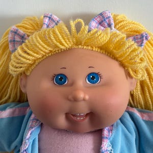Pode incluir: Boneca vintage com rosto redondo, olhos azuis e expressão sorridente. A boneca tem cabelo amarelo de lã penteado com pequenos laços xadrez. Ela usa uma roupa azul claro e rosa.