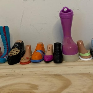 Può includere: Una collezione di 20 scarpe di plastica colorate per bambole, tra cui stivali, sandali e scarpe da ginnastica. Le scarpe sono di vari colori, tra cui rosa, blu, rosso, verde e giallo.