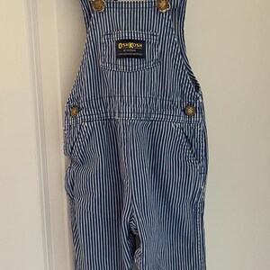Puede incluir: Pantal&oacute;n vaquero azul y blanco a rayas con un parche de logotipo OshKosh B'Gosh dorado. El pantal&oacute;n vaquero tiene tirantes ajustables con hebillas doradas.