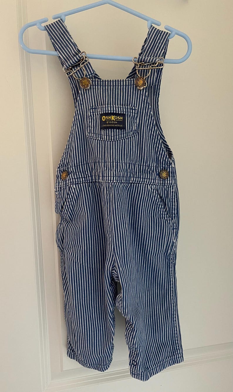 Puede incluir: Pantal&oacute;n vaquero azul y blanco a rayas con un parche de logotipo OshKosh B'Gosh dorado. El pantal&oacute;n vaquero tiene tirantes ajustables con hebillas doradas.