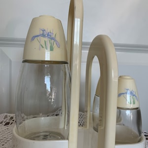 May include: White ceramic condiment holder with three glass bottles. The bottles have white lids with a floral design of purple irises.