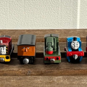 May include: A row of toy train engines in various colours, including blue, green, and red, are displayed on a wooden board. Each train has a distinct design and facial expression, representing different characters.