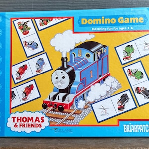 May include: A blue box featuring a Thomas the Tank Engine domino game. The box has a blue border and a yellow background with dominoes and a cartoon train. Text on the box reads "Domino Game" and "Thomas & Friends".