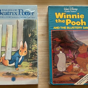 May include: Two vintage VHS tapes. One features "Tales of Beatrix Potter" with a rabbit illustration. The other is "Winnie the Pooh and the Blustery Day" with characters from the animated film. Both tapes are in plastic wrap.