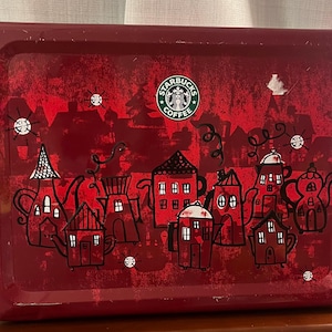 May include: Red metal tin featuring a Starbucks Coffee logo and a whimsical illustration of houses with chimneys and roofs. The houses are arranged in a row and have a cartoonish style.