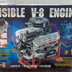 Puede incluir: Un kit de modelo de motor V-8 Visible Revell. La caja presenta un modelo de motor de plástico transparente con componentes negros y rojos, y el texto "VISIBLE V-8 ENGINE". El kit incluye una manivela para operar el cigüeñal, los pistones y las válvulas. El modelo mide 35,56 cm.