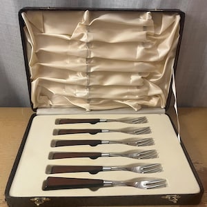 Peut inclure: Un ensemble de six fourchettes avec des dents en acier inoxydable et des manches en bois brun foncé, présentées dans un étui marron. L'étui est ouvert, révélant les fourchettes soigneusement rangées.