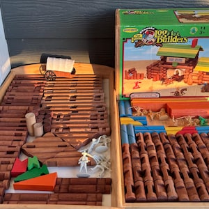 Peut inclure: Un ensemble de jouets en bois avec 170 pièces, comprenant des bûches, des planches et des figurines. La boîte montre un chalet en rondins et le texte "Classic Log Builders".