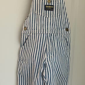 Puede incluir: Pantal&oacute;n vaquero de rayas azules y blancas con la marca OshKosh B'Gosh. El pantal&oacute;n tiene tirantes ajustables y un bolsillo delantero.