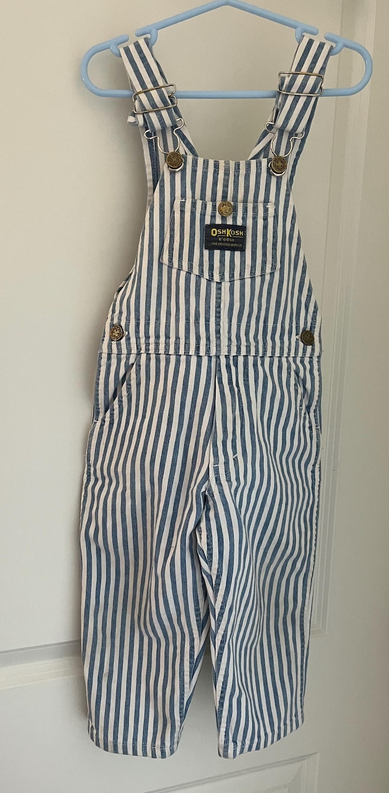 Puede incluir: Pantal&oacute;n vaquero de rayas azules y blancas con la marca OshKosh B'Gosh. El pantal&oacute;n tiene tirantes ajustables y un bolsillo delantero.