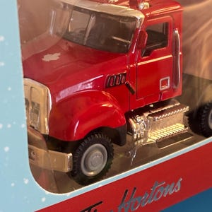 Puede incluir: Un camión semirremolque de juguete rojo con detalles plateados, exhibido en un embalaje de plástico transparente. El camión presenta un diseño detallado, incluyendo una parrilla cromada y ruedas. El embalaje incluye el logotipo de "Tim Hortons".