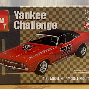 Op de afbeelding: Een 1:25 schaalmodelkit van een rode Dodge Challenger met een zwart dak en het nummer 76 aan de zijkant. De doos heeft de tekst "Yankee Challenge" en "Skill 2".