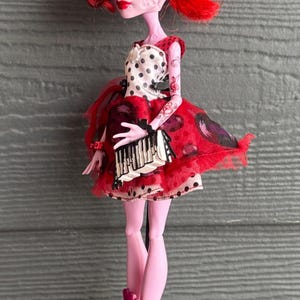 May include: A doll with bright red hair and a red and black dress. The doll has pale pink skin and is wearing a dress with a white polka dot top and a red skirt. The doll is standing on a black stand.
