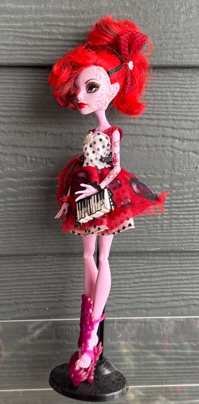 May include: A doll with bright red hair and a red and black dress. The doll has pale pink skin and is wearing a dress with a white polka dot top and a red skirt. The doll is standing on a black stand.