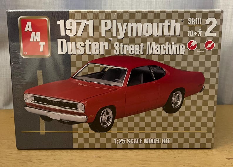 Op de afbeelding: Een modelbouwdoos voor een 1971 Plymouth Duster Street Machine. De doos heeft een rood 1971 Plymouth Duster model op een geruite achtergrond. De doos is gelabeld met "Skill 2, 10+ jaar".