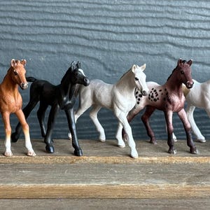 Puede incluir: Una colección de diez figuras de caballos de juguete en varios colores y patrones. Los caballos están dispuestos en fila, mostrando una gama de tonos que van del blanco y marrón al negro. Cada figura está detallada con características realistas.