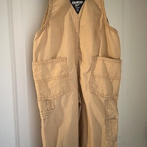 Puede incluir: Pantal&oacute;n vaquero de color beige con cuello en V y tirantes ajustables. El pantal&oacute;n tiene dos bolsillos grandes en la parte delantera y un bolsillo m&aacute;s peque&ntilde;o en la pierna derecha. El logotipo de OshKosh B'Gosh es visible en la parte delantera del pantal&oacute;n.