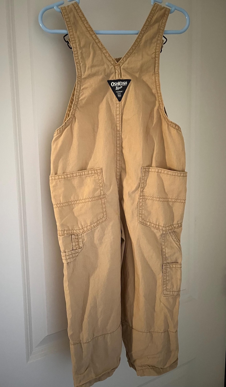 Puede incluir: Pantal&oacute;n vaquero de color beige con cuello en V y tirantes ajustables. El pantal&oacute;n tiene dos bolsillos grandes en la parte delantera y un bolsillo m&aacute;s peque&ntilde;o en la pierna derecha. El logotipo de OshKosh B'Gosh es visible en la parte delantera del pantal&oacute;n.