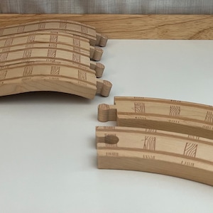 Puede incluir: Un conjunto de vías de tren de madera, que incluye piezas curvas y rectas. Las vías son de madera marrón claro y tienen un patrón a cuadros.