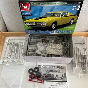 Puede incluir: Un kit de maqueta de un Dodge Charger R/T de 1971 en amarillo con rayas negras. La caja incluye las piezas del coche, las instrucciones y una hoja de calcoman&iacute;as. La caja est&aacute; etiquetada con "1971 Dodge Charger" y "Muscle Cars - Voitures &agrave; haute performance".