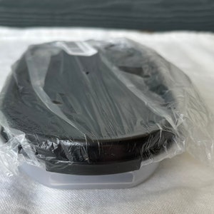 Puede incluir: Un recipiente de almacenamiento de alimentos de plástico negro con una tapa transparente. El recipiente tiene forma ovalada y una superficie lisa. El recipiente está envuelto en plástico.