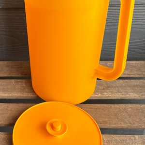 Può includere: Brocca di plastica arancione brillante con coperchio abbinato. La brocca ha un'ampia maniglia e un beccuccio per versare facilmente. Il coperchio è posizionato accanto alla brocca. La brocca è progettata per liquidi.