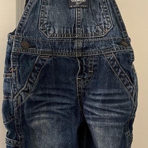 Puede incluir: Pantal&oacute;n vaquero azul con tirantes ajustables con rayas rojas y blancas. El pantal&oacute;n tiene un parche en la parte delantera que dice "OSHKOSH B'Gosh THE GENUINE ARTICLE".